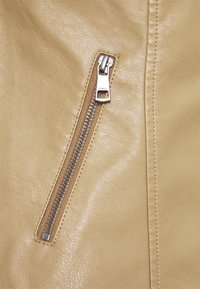 Giacca in finta pelle color beige con dettagli, caratterizzata da una tasca con zip e hardware argentato, superficie texturizzata e cuciture visibili lungo i bordi.