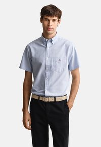 Mladý muž nosi svetlomodrú krátkoročnú košeľu s gombíkmi, béžový opasok a čierne nohavice, stojí s jednou rukou v vrecku.
