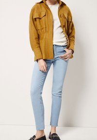 Senapsgul kordjacka med två bröstfickor, parat med ljusblå smala jeans och svarta tofflor.