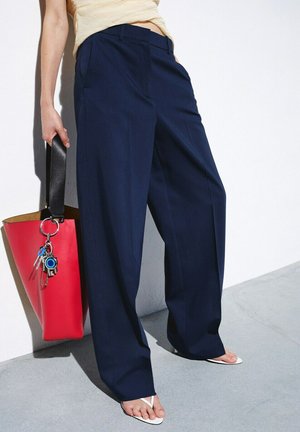 Persona con pantalones anchos azul marino y sandalias blancas tipo chancla, sosteniendo un bolso rojo con correa negra y llavero, contra una pared blanca.