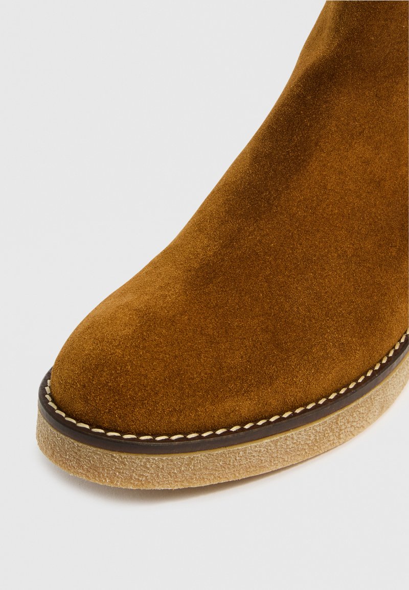 Brązowy zamszowy botek za kostkę z okrągłym noskiem, z teksturowaną cholewką i kontrastującą, jasnobeżową podeszwą z przeszyciami.