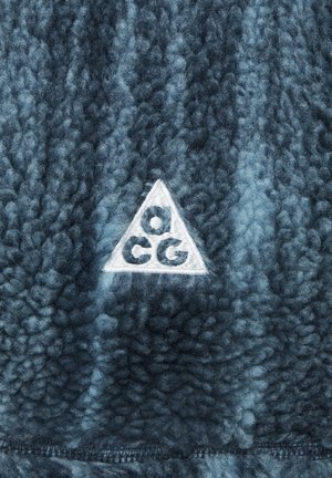Material de forro polar azul con una textura suave, que presenta un logo triangular blanco con "ACG" en su interior. La tela muestra diferentes tonos de azul.