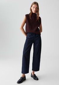 Donker bordeauxkleurige mouwloze trui met een hoge hals, gecombineerd met wijde donkerdenim jeans en zwarte instappers. Glad materiaal en casual pasvorm.