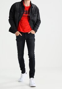 Chaqueta de mezclilla negra sobre una camiseta roja con estampado gráfico negro, combinada con jeans negros y zapatillas blancas. Diseño casual y ajustado.