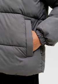 Szara pikowana kurtka puchowa z elastycznymi mankietami i bocznymi kieszeniami z widoczną metką marki na dole.