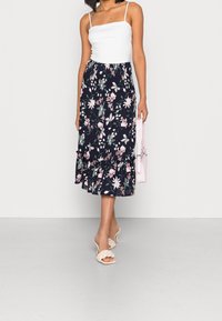 Kwiecista maxi spódnica na granatowej bazie, ozdobiona różowymi i białymi kwiatami, z warstwowymi falbanami u dołu. W połączeniu z białą bluzką i jasnym obuwiem.