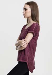 Burgundijas īsām piedurknēm tops ar V-veida kakla izgriezumu un sarullētām piedurknēm, ar smalku heather tekstūru un garāku aizmugurējo malu, kombinēts ar melnajām legingām.