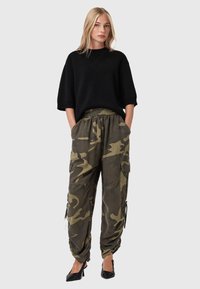 Donna bionda che indossa una maglia nera larga, pantaloni cargo mimetici e scarpe con tacco nere, in piedi con le mani nelle tasche su uno sfondo grigio.