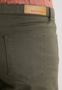 Pantalones de algodón verde oliva con un bolsillo trasero, detalles cosidos y una parche de cuero marrón claro con el texto "NAF NAF" grabado en él.