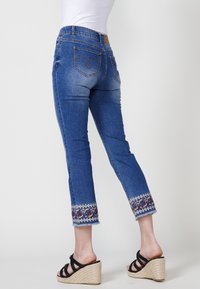 Blå denimjeans med en figurnära passform, uppvikta sömmar med ett färgglatt blommönster och slitna kanter, tillsammans med svarta klacksandaler.