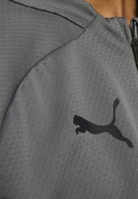 Harmaa vetoketjullinen takki, jossa on teksturoitu kangas. Rintakehässä näkyy selkeästi musta Puma-logo.