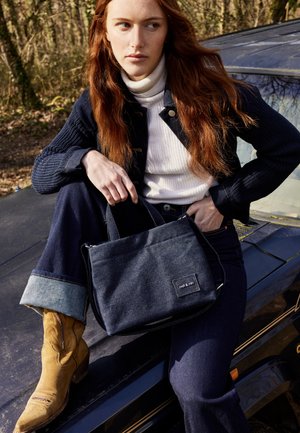 Borsa in denim di tessuto blu scuro con chiusura a zip e patch con logo. Il modello indossa un dolcevita bianco e denim scuro. Stivali da cowboy marroni.