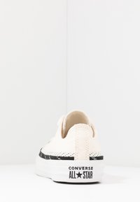 Une basket blanche Converse All Star vue de dos, montrant un détail de bande noire et le logo sur l'onglet talon de la semelle.