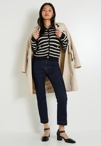 Femme portant un trench-coat beige par-dessus un cardigan rayé noir et blanc, un jean bleu foncé et des chaussures à talons blocs noires, posant contre un mur blanc.