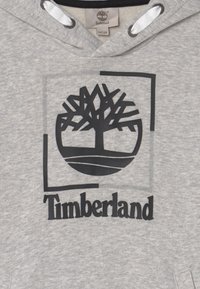 Szara bluza z kapturem z czarnym logo Timberland przedstawiającym drzewo oraz napis w obrębie prostokątnej ramki na przodzie.