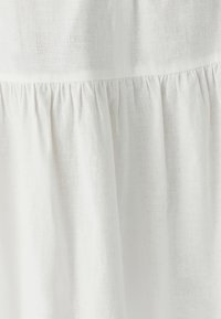 Bílé lněné šaty s texturovaným povrchem a nazbíraným detailem na lemu, který vytváří plynulý vzhled.