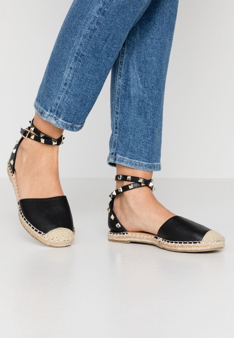 Pies que llevan espadrilles negras con correas al tobillo y tachuelas plateadas, combinados con jeans cortos azules contra un fondo blanco.