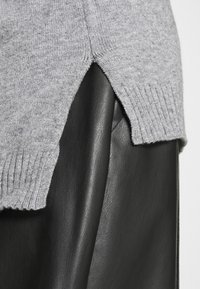Maglione grigio a maglia con orlo a costine, caratterizzato da un design con spacco laterale, indossato sopra un capo in pelle nera liscia. Contrasto di texture visibile.