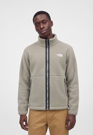 Homme portant une veste polaire gris clair zippée avec le logo The North Face et un pantalon beige sur fond blanc uni.