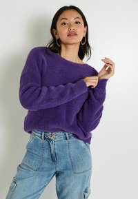 Suéter morado, esponjoso, con un escote amplio y mangas largas, combinado con unos jeans azul claro que tienen un botón y bolsillos frontales.