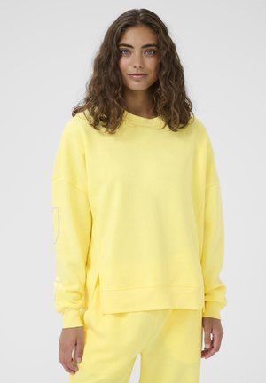 Mladá žena s vlnitými hnědými vlasy, která nosí volnou žlutou mikinu a kalhoty ve stejném odstínu, stojící před světle šedým pozadím.