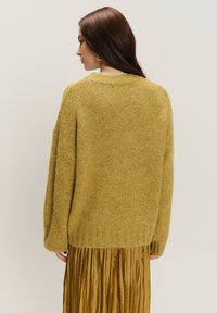 Senfgelbes, übergroßes Sweatshirt mit strukturiertem Strick, geripptem Saum und überschnittenen Schultern, kombiniert mit einem plissierten goldenen Rock.