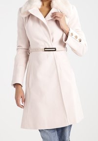 Manteau rose clair avec un col en fausse fourrure, taille ceinturée et manches longues. Doté d'un tissu texturé et de boutons dorés aux poignets.