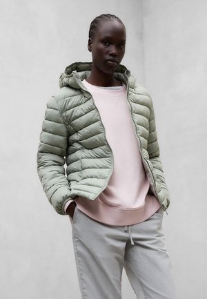 Mujer con chaqueta acolchada verde claro sobre una sudadera rosa pálido y pantalones gris claro, de pie con las manos en los bolsillos contra un fondo liso.