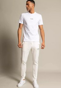 Biała koszulka z krótkim rękawem z logo z przodu, zestawiona z białymi spodniami dresowymi z bocznymi paskami i logo na kieszeni. Białe trampki.