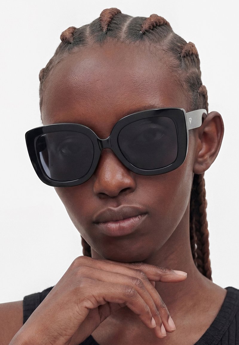 Femme à la peau foncée et aux cheveux tressés porte de grandes lunettes de soleil noires, reposant son menton sur sa main avec une expression neutre.