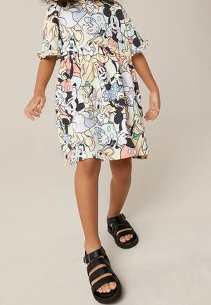 Enfant portant une robe longueur genou avec des personnages Disney et des sandales noires à boucle, se tenant sur un fond neutre.