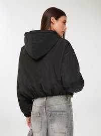 Giacca nera con cappuccio realizzata in un materiale lucido e leggero. Caratterizzata da un design voluminoso con orlo elasticizzato, abbinata a jeans grigi chiari sbiaditi.