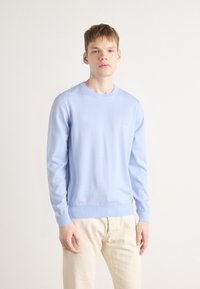 Suéter azul-claro de gola redonda feito de um tecido suave, com mangas longas e bainha canelada. Combinado com calças de cor creme.