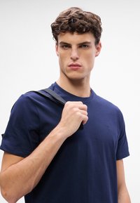 Jeune homme aux cheveux courts, bouclés et bruns, portant un t-shirt bleu marine, tenant une sangle de sac noire sur son épaule sur fond blanc.