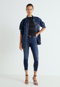Chaqueta de mezclilla y top negro ajustado combinados con jeans oscuros ajustados. Incluye detalles de desgaste sutil, botones clásicos y tacones con tiras.