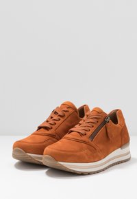 Par av rostorange mockaskor med snörning, sidodragkedja och vita randiga sulor på vit yta med en enkel grå bakgrund.