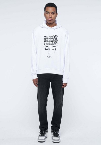 Felpa bianca con stampa grafica che presenta testo e un viso, abbinata a jeans scuri e sneakers. Tessuto liscio, vestibilità casual.