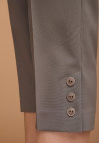 Graue verkürzte Hosen mit seitlichem Schlitz und drei kleinen braunen Knöpfen am Saum, aus glattem Stoff gefertigt.