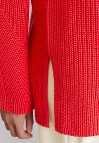 Gros plan sur une personne portant un pull en tricot rouge avec une texture côtelée et une fente sur le côté, associé à un pantalon de couleur claire, la main décontractée sur le côté.