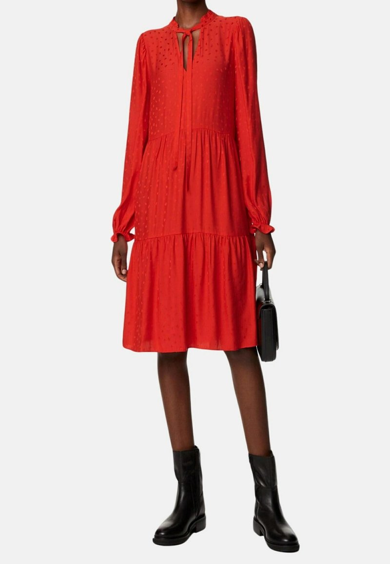 Robe rouge avec un design à niveaux, longues manches bouffantes et un col à lacets. Présente des motifs en relief à pois. Portée avec des bottines noires.