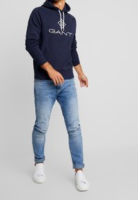 Felpa navy con logo bianco "GANT" sul petto, tasca a canguro, cappuccio con cordino, abbinata a jeans azzurri e sneakers bianche.