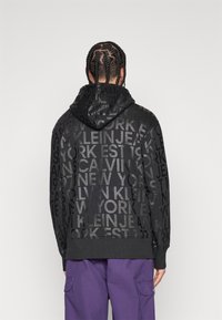 Black hoodie featuring a textured repeating text pattern in gray. It has a drawstring hood and ribbed cuffs, with a solid black lower section.