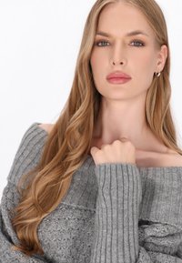 Grå off-shoulder tröja med ribbade ärmar och en texturerad kropp. Mjukt stickat material med lång päls som ramar in ansiktet. Enkla silvriga örhängen.