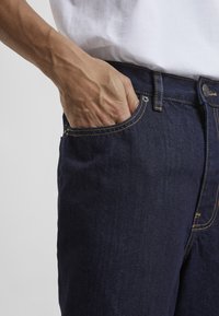 Tummansiniset farkut, joissa on klassinen suora leikkaus, kuparinväriset tikkausyksityiskohdat ja etutasku, johon käsi lepää.