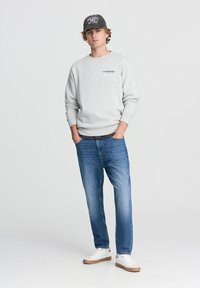 Felpa grigia in pile con polsini a coste, abbinata a jeans a gamba dritta blu chiaro. Sneakers bianche con suola beige e un cappellino grigio con testo.