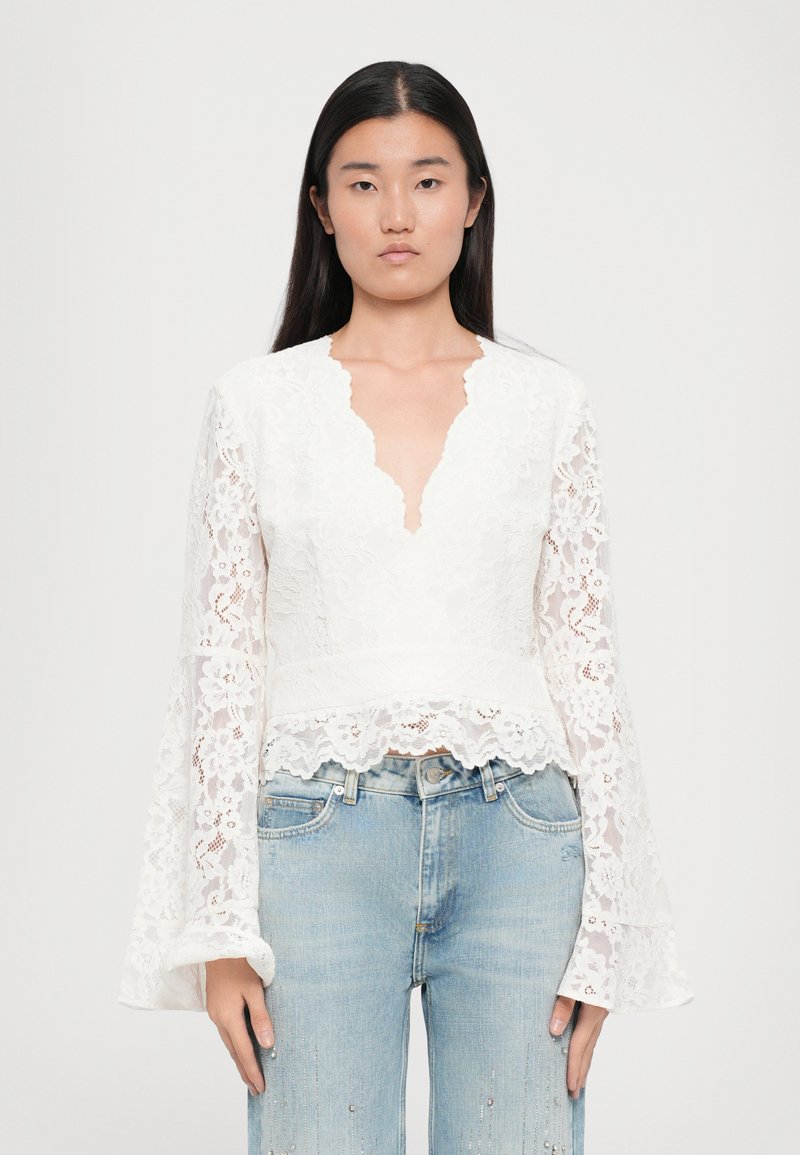 Witte kanten blouse met een V-hals, lange belmouwen, geschulpte zoom en bloemenpatronen, gecombineerd met lichtblauwe jeans.