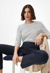 Gestreiftes longsleeve Hemd in Weiß und Marineblau, kombiniert mit marineblauen High-Waist-Hosen. Das Model sitzt mit einer hellbraunen Jacke, die in der Nähe drapiert ist.