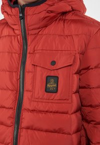 Giacca puffer rossa dal finish lucido, con chiusura a zip frontale, cappuccio e una tasca sul petto con un dettaglio del logo a patch nero.