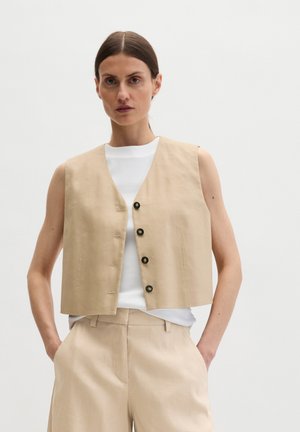 Vrouw draagt een beige mouwloos gilet met knopen over een wit topje en bijpassende beige broek, staat met handen in de zakken tegen een witte achtergrond.