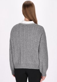 Pulover gri din tricot cu tiv ribbut și textură împletită în cablu. Are o croială lejeră și mâneci largi, purtat peste o cămașă albă cu guler.